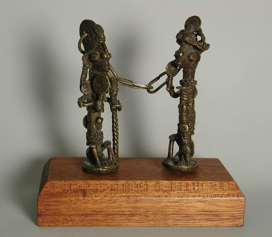 Hearst Museum object titled Figures, accession number 5-16317, described as Ogboni society figures; pair of Ogboni society figures connected by a chain and mounted on a wooden block, cast brass figures, (a) 1 male, (b) 1 female, each holding a staff and rattle. both have elaborate hair treatment and head pieces