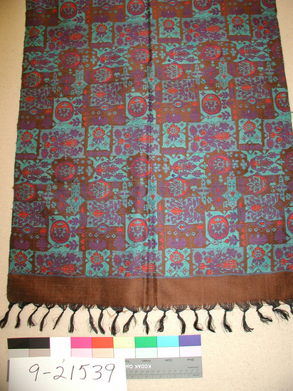 Hearst Museum object titled Shawl, accession number 9-21539, described as Shawl; W=57 cm x L=185 cm; 6.0 cm warp fringe knotted once; black warp 5 cm borders on each end with bronze-like weft; bronze field printed in palmettes, flowers in vases, stylized crosses and random smaller floral and geometric motifs and turquoise, purple, red, orange scattered over the field; tied warp fringe in black; dye on back comes out white, red and light blue