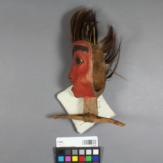 Hearst Museum object 3 of 6 titled Headdress ornament, accession number 2-19098, described as In form of small mask representing human head, face red on one side, green on the other, human hair and duck quill fringe. Mounted on carved, broken wood rod. Upper portion of larger piece.