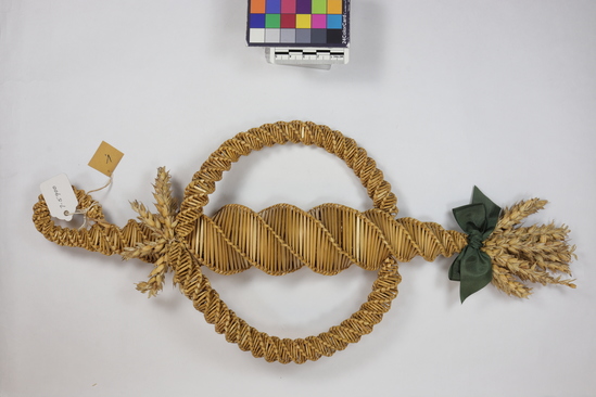 Hearst Museum object titled Corn dolly, accession number 7-5900, described as corn dolly; wheat straw, plaited; shaped like ear of corn with stem end bent over to form loop; tassel of wheat heads at other end, tied with green ribbon bow; plaited circle attached perpendicularly, trimmed with wheat heads at top; 1.44 centimeter; maximum width 19 centimeters.