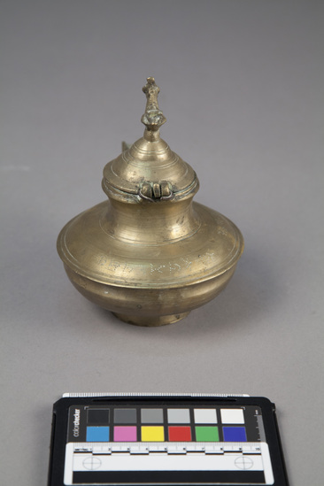 Hearst Museum object titled Vessel, accession number 9-12916, described as Cast bronze vessel with a spout, a hinged lid with a peacock finial (broken tail); cast grass.  Native name and meaning: "varhi" a vessel for ghee. Used by Hindu's for pouring ghee, either in religious or domestic contexts. A saying in Gujerati "ghee varhi ma thi pissar_ju {partially illegible} or "ghee flowed from a varhi", signified lavish entertainment, because the ghee is poured without lifting the spout between place-settings. In its household context, this vessel is used for storing and pouring ghee. A vessel of this type is used in the worship of Vishnu by followers of Vallabhacharya, the founder of the Pushti Marg cult, who believe that the deity drinks water from it in their absence. Height approximately 16 cm. (including the finial).