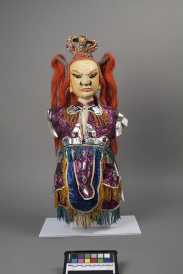 Hearst Museum object 6 of 6 titled Hand puppet, accession number 9-22416, described as Puppet, hand. King.   A.  Head:  carved wood, off-white with orange, green, brown and black features, red-orange hair with 2 high ponytails, crown:  wire wrapped in fabric with pink, blue white and gold beads  B.  Body: white cloth.  Legs:  green-brown cloth.  Hands: wood, white, orange nails.  Boots:  wood, black and white.  C. Robe:  cloth,  metallic purple, silver applique, yellow and blue fringe.  51 x 24 cm (20 1/16 x 9 7/16 in)