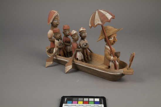 Hearst Museum object 5 of 15 titled Figures, accession number 5-16237a-q, described as caricature figure group; caricature of a District Officer on tour; carved & painted wood a - boat, has bag of frags b - Dist. officer (front) c - poler (rear) d - paddler (LF) e - paddler (LR) f - paddler (RF) g - paddler (RR) h - hat i - umbrella handle j - umbrella k - flag l - pipe m - pole n - paddle (RF) o - paddle (LF) p - paddle (LR) q - paddle (RR) Found two pegs in a bag with this object number.