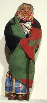 Hearst Museum object titled Doll, accession number 2-71784, described as Cochiti Pueblo Indian woman doll. Cotton stuffed doll in costume of Cochiti Pueblo. Dressed in one should dress of black fabric, Black yarn hair, molded and painted features. White leather leggings and shoes. Rayon satin shawl with embroidered lining. Height 11.5 inches