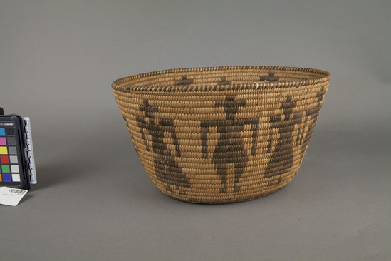 Hearst Museum object 3 of 3 titled Basket, accession number TB-94, described as Per Ralph Shanks:  Tourist trade basket.  Possibly acc. 1103 (first number hard to read) on tag. Oval coiled basket with cattail bundle foundation.  Black designs are probably yucca.  Alternating human figures: 5 men and 5 women both wearing hats. Rim ticks/striped.  Coiled with clipped weft fag ends.