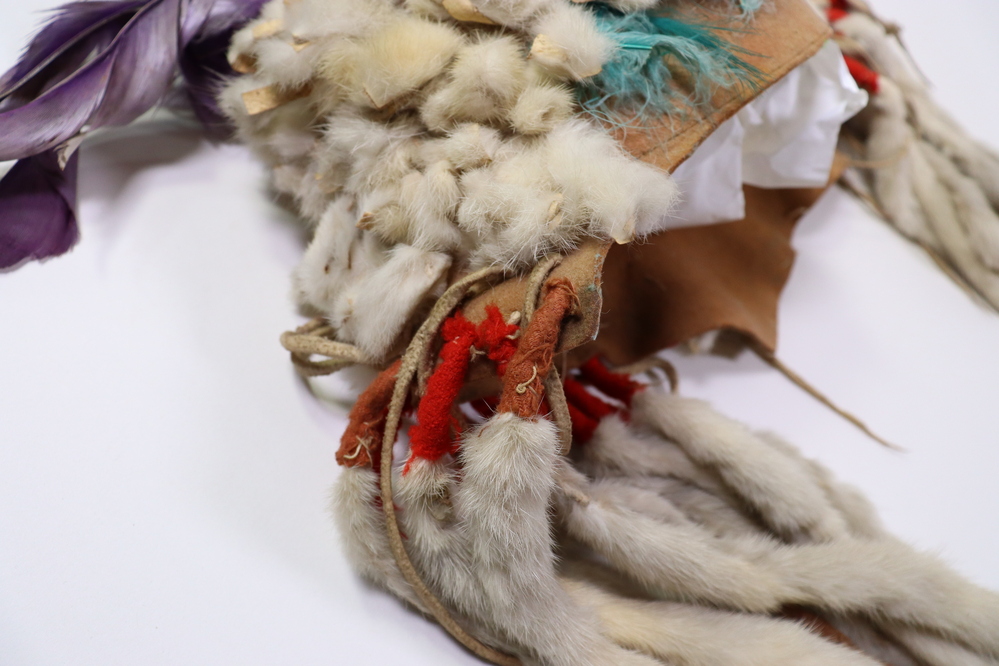 Hearst Museum object 10 of 15 titled Headdress, accession number 2-45410, described as Felt cap covered with strips of rabbit fur, patches stained yellow, traces of green fluff at front. Crown with 2 split polished horns attached to interior hide strip. Purple feathers and green-gray fluff in between and at base of horns. Multicolored silk grosgrain (?) ribbons bound to left horn tip. Ten "ermine tails" of rabbit fur sewn about a cord, red felt seized, with black hair tips; fringes at sides.