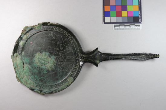 Hearst Museum object 8 of 13 titled Stemmed mirror, accession number 8-5859, described as Etruscan bronze mirror; cast-handle type; encrustation on both surfaces of disk and blue discoloration on obverse due to bronze disease; dark green patina; handle ends in stylized ram’s head; rim of obverse beaded; engraved scene of four figures, two women flanking Minerva (Greek=Athena) and nude man, standing against architecture; guilloche border; mirror appears to have been conserved with a translucent glaze.