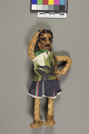 Hearst Museum object titled Woven reed doll, accession number 5-14452, described as Doll, female, plaited palm leaf, with brown yarn embroidered features and hair. Wearing multicolored bead embroidered skirt, with a green print blouse. Height 18.2 centimeters.