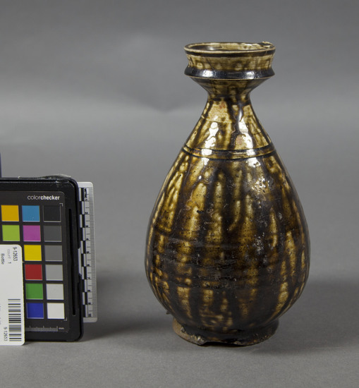 Hearst Museum object titled Bottle, accession number 9-12653, described as Oil bottle; stoneware, tear-drop shape, wide mouth rim on constricted neck, narrow foot covered with coarse sand particles, chip out of foot, shiny glaze that is rust brown where thick, green where thin, slip (?) underneath glaze, two incised bands on upper body, 4 7/8” (11.8 cm) diameter, 8 5/8” (21.8 cm) high.