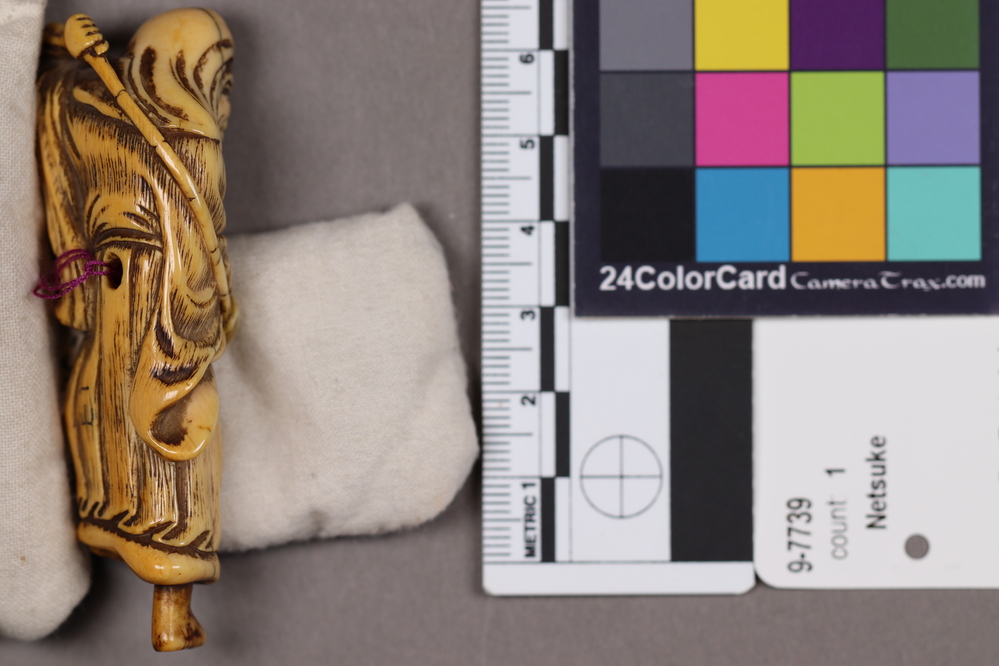 Hearst Museum object 7 of 9 titled Netsuke, accession number 9-7739, described as Netsuke: tall thin man with long face. Left foot raised, holds long curved rod behind his back.