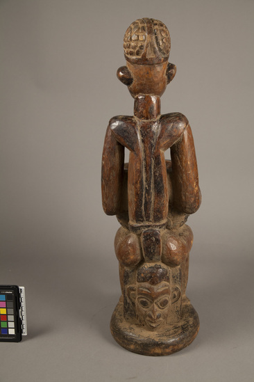 Hearst Museum object 4 of 4 titled Carved wood figure, accession number 5-6001, described as Wood male figure seated on stool; holding bowl with human-like figures in low relief; stool is carved human head; h. 63.5 cm. Fine brown patina; some termite damage on base.