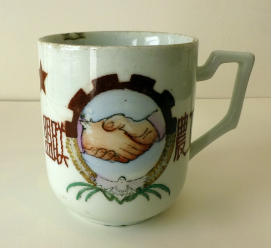 Hearst Museum object titled Mug, accession number 9-23483, described as Distinctive 1950s angular handle. The hand-painted decal shows two hands shaking are encircled by the ring of a cog, a dove of peace and friendship at the bottom. Two characters on each side, read from right to left: “Worker-Peasant Alliance” (工農联盟 Gongnong lianmeng). A five-pointed star at upper left.  Inscription: “Energetically produce” (努力生產 Nuli shengchan). Kiln mark: “Product of Liling Ceramic Painting Worker’s Commune” (醴陵陶畫工人合作社出品Liling taohua gongren hezuoshe chupin)