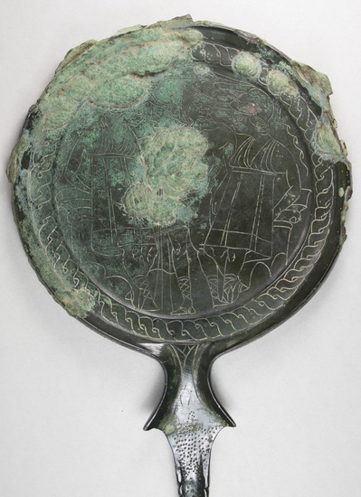 Hearst Museum object 4 of 13 titled Stemmed mirror, accession number 8-5859, described as Etruscan bronze mirror; cast-handle type; encrustation on both surfaces of disk and blue discoloration on obverse due to bronze disease; dark green patina; handle ends in stylized ram’s head; rim of obverse beaded; engraved scene of four figures, two women flanking Minerva (Greek=Athena) and nude man, standing against architecture; guilloche border; mirror appears to have been conserved with a translucent glaze.