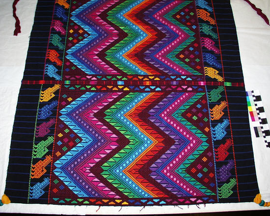 Hearst Museum object 1 of 2 titled Tzute, accession number 3-29959, described as Backstrap-loomed, warp-predominant plain weave; two-faced supp. weft brocading; two  pieces joined with randa; one end loom-finished, other end warps cut and hand-hemmed. 11" tassels added to all four corners. 96.5 cm x 96.5 cm. Native name/meaning: Tzute, hombre/Man's headcloth. Materials, techniques: Cotton: 2 two-ply black, blue and 4 two-ply black; mercerzied cotton in 5 two-ply in multicolors. Iconography consists of animals, abstracted double eagles, in geometrics. Context of use: Donated by Brian Tapp in memory of Rodolfo Alvarez Farfan. Zigzag patterns are an abstract rendering of the double-headed eagle design (Schevill); handsome tassels at four corners begin with yellow around which is an aqua puffed ring; the yellow is then knotted to maroon cotton, the dominant color of the tassel. The tassels are each knotted towards the bottom.