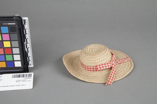 Hearst Museum object 3 of 3 titled Doll, hat, accession number 5-15759a,b, described as (a) Rag doll; black woman or child, wearing white blouse and checked red skirt; ring on finger of left hand; painted facial features and black hair. Height ca. 43.0 cm. (b) Hat, raffia; coiled bands sewn together; rounded crown and flat brim with hat band of same material as skirt. Diameter 15.5 cm. Made in craft classes in the schools and sold at Home Industries.