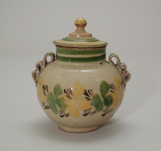 Hearst Museum object titled Jar with lid, accession number 3-29466a,b, described as jar, covered; earthenware with lead glaze; (a) jar: globular with folded clay self handles on both sides; green, yellow and brown abstracted floral design band on buff; two encircling green bands around neck; height 12.9 cm; (b) cover, round with self finial; encircling yellow and green bands; rim diameter 8.9 cm. Purchased at Berkeley antique shop for Lowie Museum of Anthropology by Barbara E. Busch - University Appropriation.