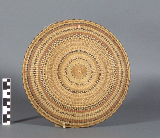 Hearst Museum object titled Plaque, accession number 10-1083, described as Basketry plaque; twined and wicker; circular; round bottom; shades of brown, natural; scroll design around edge; 13 3/8”dia.