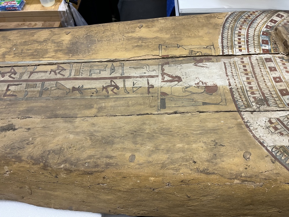 Hearst Museum object 103 of 167 titled Coffin, accession number 6-19912, described as Wooden anthropoid coffin. Inscription running down center of lid. Determined by Kea Johnston to probably be the outer coffin of Iwefaa (6-19928, 12-11039). According to Kea Johnston, "This coffin belonged to a man named Iwefaa, whose name is inserted in the middle of the spells written in the two columns on the front. This coffin is likely the outer coffin for PAHMA 6-19928 based on the similarity of the owner's name, unusual costume, and the paleography of the inscription. Both coffins were purchased on the antiquities market by George Reisner for Phoebe A Hearst. However, stylistically, they are very similar to a set of coffins known to have come from the site of Akhmim, which was excavated in the 1880s by Gaston Maspero, and which has heavily looted for the antiquities market during this decade." "The pectoral vignette between the text and the collar shows the deceased standing before squatting baboon and is the source of our logo. The hieroglyph for 'sky' is above their heads, and the side borders are formed by a pyramid and the symbol for the West.