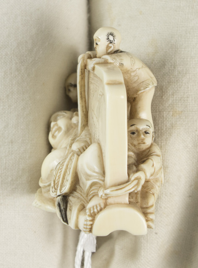 Hearst Museum object 2 of 14 titled Netsuke, accession number 9-18870, described as Netsuke; reclining figure of fat man facing to his right, supporting head on right hand; lying in front of wall; boy at left peering at man; boy at back of wall standing on barrel pulling at kimono cord; boy at right tugging at kimono hem; man holding fan in left hand; both sides of wall visible; H 3.6 cm X L 4.8 cm