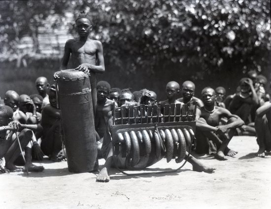 Hearst Museum object titled Black and white negative, accession number 15-32373, described as 4" x 5" Negative. One man from Kiya Be stands with a drum and another musician next to him sits in front of his instrument. A crowd sits behind them.