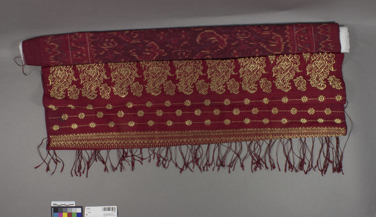 Hearst Museum object titled Shawl, accession number 7-10193, described as From Cat Card:  Stole, claret red silk, self fringe plied z twist 26cm at each end. 67cm gold supp weft design - widely spaced stripes, stripe of dense design, stripes but with flowers added (on one end emphasized by using thicker weft ground), then 12.5cm large repeated motif (tree of life or paisley like), then 5.5cm dense geometric pattern. Center is weft ikat with yellow, green, blue, black. A few darns in the ikat area - may be original. Looks like Thai silk. c1925. Cat Card Notes indicate shawl may be from Thailand? possibly Indonesia? or constructed from Asian materials?