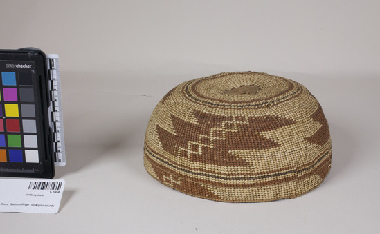 Hearst Museum object 2 of 2 titled Cap, accession number 1-1804, described as Cap basket; twined. Warp is Hazel (Corylus cornuta californica) weft is Sugar pine root (Pinus lambertiana). White pattern is Bear grass (Xerophyllum tenax). Red dye is Chain fern (Woodwardia spinulosa) in Alder (Alnus rubra). Green is mud dyed Bear grass (?). height: 7.3 cm, diameter: 18.9 cm.