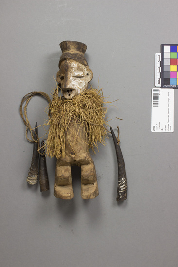 Hearst Museum object titled Figurine, accession number 5-5326, described as Wooden figurine. Torso wrapped with raffia fringe. 3 antelope/gazelle horns dangle from rope around neck. 24.6 cm