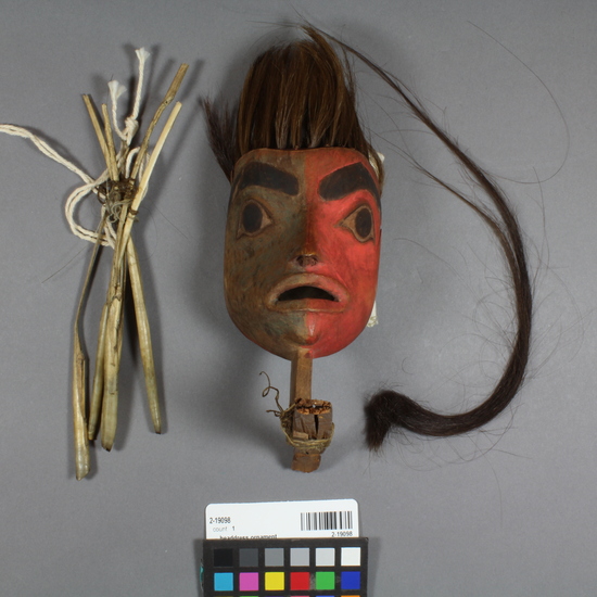 Hearst Museum object 4 of 6 titled Headdress ornament, accession number 2-19098, described as In form of small mask representing human head, face red on one side, green on the other, human hair and duck quill fringe. Mounted on carved, broken wood rod. Upper portion of larger piece.