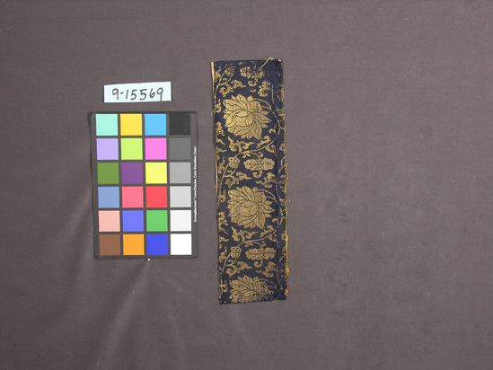 Hearst Museum object titled Textile, accession number 9-15569, described as Lined strip; silk, flat gilt paper strips, cotton lining; satin, brocaded, plain weave lining; dark blue ground, gold designs, light brown lining; lotus motives. Same as original catalog #453, 454, 455. 5 in. x 18 1/2 in.