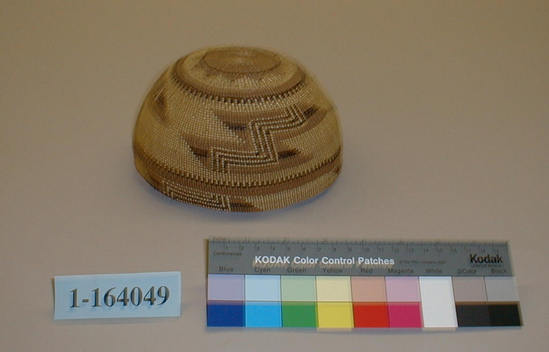 Hearst Museum object titled Cap, accession number 1-164049, described as Basketry, twined geometric designs in overlay technique; brown and yellow.  Plain-twined, peeled hazel or willow shoot warps, pine root weft overlay of beargrass leaf and maidenhair fern stems. Label: "Klamath River Tribes". Per Ralph Shanks:  Woman’s twined basket hat.  Crossed warp starting knot.  The warp material is probably hazel or willow.  The background wefts are conifer root.  The weft overlay is beargrass and maidenhair fern.  Starting at the starting knot is 1/2 inch of three strand twining, followed by 1.25 inches of plain twining.  Plain twining continues until 3/4 inch below the rim where there is another weft row of three strand twining.  After that plain twining continues to the end of the rim, which is trimmed.  The background overlay is beargrass.  Starting at the starting knot there is a 1/2 an inch of conifer root with no overlay.  That is followed by one weft row of maiden hair fern.  A pattern of a sun/flower in conifer root is made by an outline of beargrass, with one weft of maidenhair fern at the tip of each the rays.  The main design is made of three elements comprised all three materials that include two stacked parallelograms with a zigzag through it.  The basket has an up to the right slant of weft twist.  The workface is on the exterior, with a rightward work direction.  The overlay is only on the exterior.  The basket is from Northwestern California.