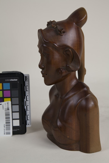 Hearst Museum object 7 of 8 titled Bust, accession number TEMP 2018.998, described as carved wooden female bust (head) signed Sinpar. 9.5"h with long hair pinned up; she wears a headdress, earrings, and strapless garment with cloth over her left shoulder; bust stops at waist and elbows; on bottom is carved "Io Bc. Kp SIMPAR, SANUR., BALI."; "6250" written in black pen on bottom.