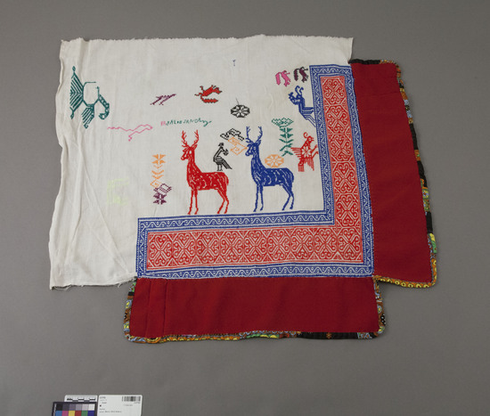 Hearst Museum object titled Shawl, accession number 3-27783, described as Cape; white gauze-like cotton square partially bordered on two sides with red synthetic material; two deer (one red, one blue), two birds, other designs in varicolored cross-stitch embroidery; signed "Bashlao (Basilio) Sanchez"; embroidered curvilinear and geometric border in red and blue edges on edges adjacent to red material; overall 78 x 75.5 cm.