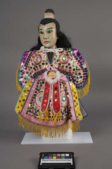 Hearst Museum object 9 of 10 titled Hand puppet, accession number 9-22260, described as Puppet, hand.  Young Man.  A.  Head:  wood, carved, white and pink with green, pink and black features, black hair in bun.  B.  Body:  cloth, brown.  Legs:  cloth, green and pink.  Hands:  wood, white.  Boots:  wood, black and white.  C. Robe:  cloth, pink with green, pink and gold sequins.  yellow fringe.  50 x 36 cm (19 11/16 x 14 3/16 in).