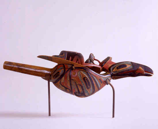 Hearst Museum object titled Rattle, accession number 2-19094, described as Totemic wooden rattle in raven form, human being on back with frog on his stomach and hawk head behind his legs, raven "ears" connect with head of human figure, sparrow hawk carving on underside; alder; painted red, blue, and black. Frog sitting on human figure is sticking its tongue into mouth of human. Raven head with frog in mouth refers to a myth where raven took frog in its mouth and descended to the depths of the sea, frog acting as lungs for raven. Raven, frog and wood identified by Charles Brown.