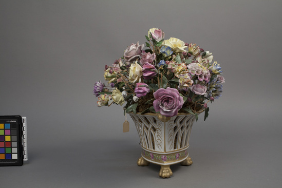 Hearst Museum object titled Decorative flower arrangement, accession number 7-7732, described as Container with flowers: porcelain, moss, wire.  Pierced container with four feet, stuffed with moss holding many porcelain flowers on wires.  White ground, gilt, green, red designs; red, yellow, blue flowers 11" overall height, approximately.  Victorian era.