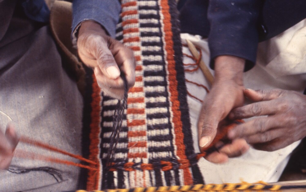 Hearst Museum object titled Color slide, accession number 25-24950, described as 35mm color slide transparency: "Najrani men weavers twining". 2 people's hands weaving a red, black, and tan colored striped textile.