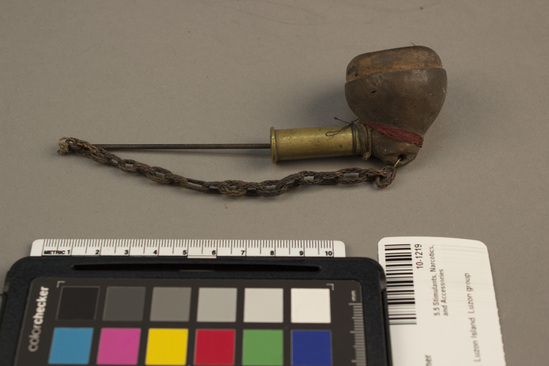 Hearst Museum object 2 of 2 titled Pipe with cleaner, accession number 10-1219, described as Pipe with wooden bowl and attached needle on chain of braided rattan; short stem piece is part of cartridge shell; bowl 1 1/2” dia. Stem missing, 1963.