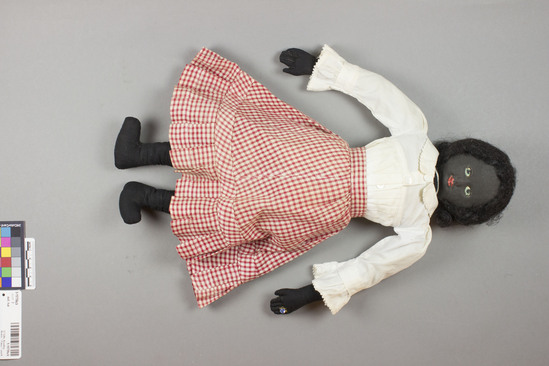 Hearst Museum object 2 of 3 titled Doll, hat, accession number 5-15759a,b, described as (a) Rag doll; black woman or child, wearing white blouse and checked red skirt; ring on finger of left hand; painted facial features and black hair. Height ca. 43.0 cm. (b) Hat, raffia; coiled bands sewn together; rounded crown and flat brim with hat band of same material as skirt. Diameter 15.5 cm. Made in craft classes in the schools and sold at Home Industries.
