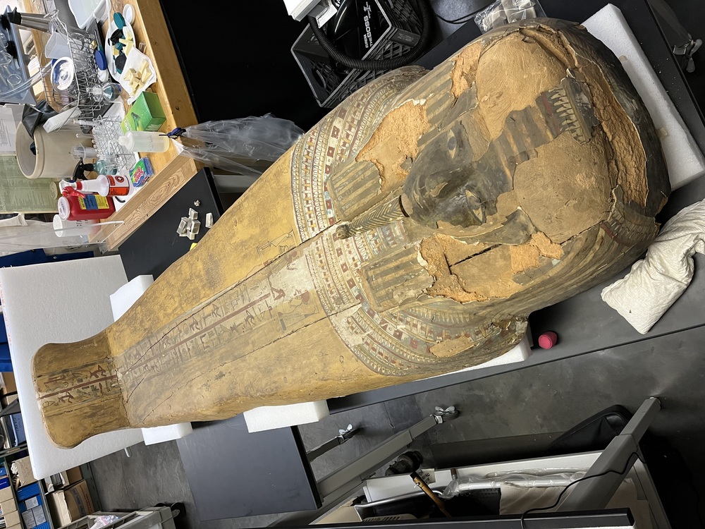 Hearst Museum object 115 of 167 titled Coffin, accession number 6-19912, described as Wooden anthropoid coffin. Inscription running down center of lid. Determined by Kea Johnston to probably be the outer coffin of Iwefaa (6-19928, 12-11039). According to Kea Johnston, "This coffin belonged to a man named Iwefaa, whose name is inserted in the middle of the spells written in the two columns on the front. This coffin is likely the outer coffin for PAHMA 6-19928 based on the similarity of the owner's name, unusual costume, and the paleography of the inscription. Both coffins were purchased on the antiquities market by George Reisner for Phoebe A Hearst. However, stylistically, they are very similar to a set of coffins known to have come from the site of Akhmim, which was excavated in the 1880s by Gaston Maspero, and which has heavily looted for the antiquities market during this decade." "The pectoral vignette between the text and the collar shows the deceased standing before squatting baboon and is the source of our logo. The hieroglyph for 'sky' is above their heads, and the side borders are formed by a pyramid and the symbol for the West.