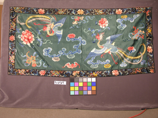 Hearst Museum object titled Hanging, accession number 9-16469, described as Hanging, embroidered silk; teal silk satin front & orange patterned silk backing, border has black ground w/ floral design interspersed w/ various objects such as fans & swords.  Main panel has 2 phoenix in flight w/ flower stem in beaks, too-1 head & cloud design & bat in background.  Silk satin w/ multicolor embroidery, mostly in satin & stem stitch but also employed french knot & a bit of couching.  Feng-hwang = phoenix.  China att.  160 cm by 76 cm.