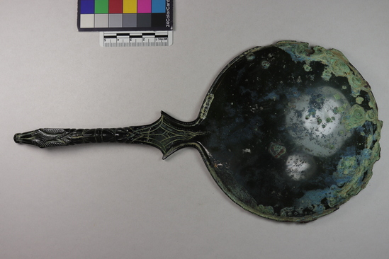 Hearst Museum object 11 of 13 titled Stemmed mirror, accession number 8-5859, described as Etruscan bronze mirror; cast-handle type; encrustation on both surfaces of disk and blue discoloration on obverse due to bronze disease; dark green patina; handle ends in stylized ram’s head; rim of obverse beaded; engraved scene of four figures, two women flanking Minerva (Greek=Athena) and nude man, standing against architecture; guilloche border; mirror appears to have been conserved with a translucent glaze.