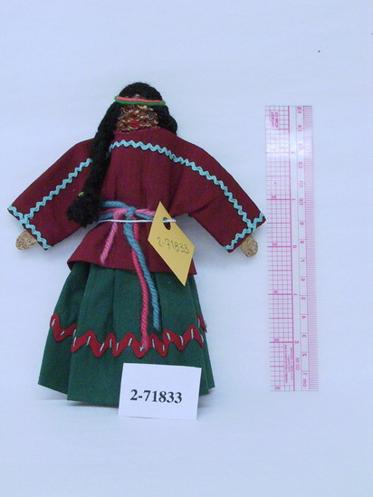 Hearst Museum object titled Female doll, accession number 2-71833, described as Corn cob doll w/ cork hands. Nails for eyes, yarn hair in braids, fabric mouth & clothing. Dressed in burgundy blouse and green skirt, both decorated with ric-rac. Yarn belt and headband.   H = 7.5”.