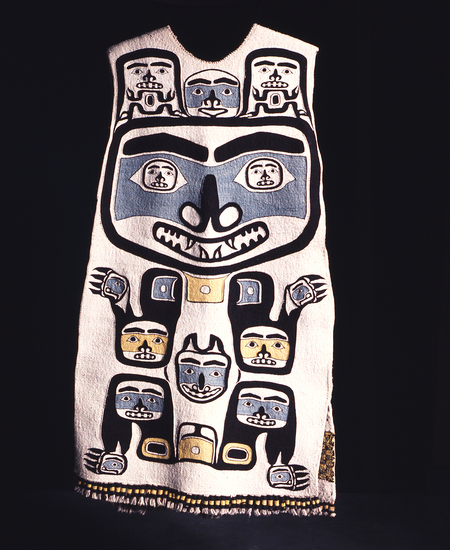 Hearst Museum object titled Tunic, accession number 2-59550, described as Abstracted bear designs; black, yellow and blue on white; dyed mountain goat wool twined over wool-covered cedar bark warp; front design is standing bear with large face; smaller faces within ears and at joints; on back are (top to bottom) a pair of eyes, 4 spinal vertebrae?, and black/yellow checkerboard panel; tassels at base of garment; one side of tunic is sewn almost closed, other side has 3 pair buckskin ties; drawstring at neck. Designs said to be grizzly bear and 2 cubs.