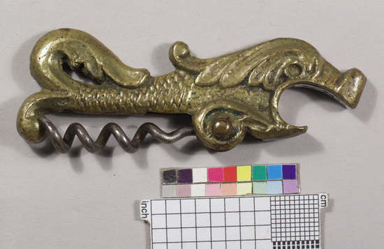 Hearst Museum object titled Corkscrew & bottle opener, accession number 7-9442, described as Corkscrew/caplifter combination, zoomorphic; cast brass tee handle in shape of sea monster, crown caplifter as mouth; iron wire helical worm pivoting on rivet near fish’s belly; 7.7 cm. long England attributed. Cast brass, iron, die forming Wine bottle cork puller and caplifter