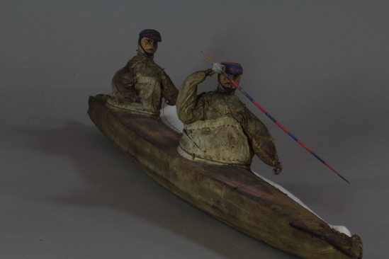 Hearst Museum object 2 of 8 titled Kayak model, accession number 2-4569, described as 2-hatch. Wood frame, rawhide cover. 2 human figures wearing hats painted bright blue and red and wearing sealskin parkas. 15 fixtures including weapons and paddles painted bright red and blue.