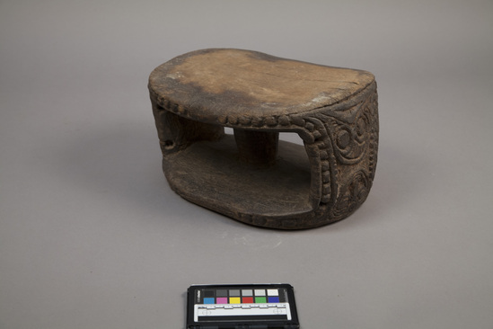 Hearst Museum object titled Stool (seating furniture), accession number 11-37713, described as stool, man’s; wood, carved relief faces at ends, oval top and bottom, columnar separator in middle; four perforations on either end; traces of pink-red pigment