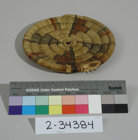 Hearst Museum object titled Plaque, accession number 2-34384, described as Basketry plaque, coiled.  Black, red, yellow, and green.  Design:  4 radii emanating from center but not connected or touching at center.  Alternating green-black, then red-black; green-yellow-red rectangular elements between each pair of radii.  Center unravelling:  hole in center.  Fibers missing in places, colors fading.