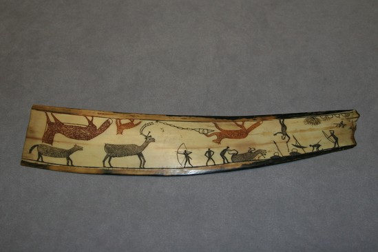 Hearst Museum object titled Carving, accession number 2-195, described as Scene on flat curving piece of ivory; umiak, seals, birds, fish seine with men, etc. incised and darkened on one side; on the other side are caribou hunters in black and red fox-like animals.