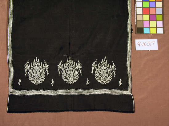 Hearst Museum object titled Shawl, accession number 9-16517, described as Shawl, black wool with blue embroidery along edges; embroidered floral designs and short fringe on each end; reverse side lined near ends; 6 circular iron-on patchers.  Wool.  India, Kashmir.  Length 170 cm.