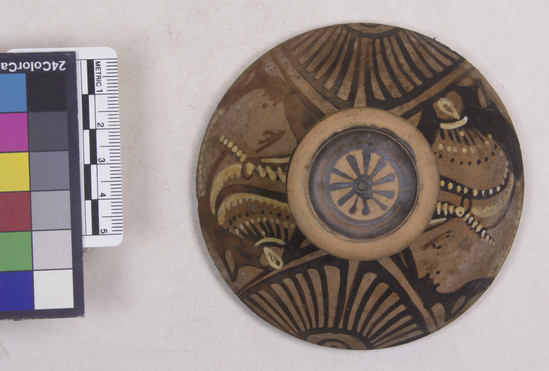 Hearst Museum object 12 of 14 titled Pyxis lid, accession number 8-12a, described as Apulian red figure pyxis lid; several small chips in rim and one in knob; slightly reddened clay; knob glazed with upper edge and central medallion, decorated with rosette, reserved; A-B, head of woman to left, wearing sakkos, diadem, earring, and neckalce; details in black and yellow; between heads, palmettes; strokes on rim. Once in Brussels in the Somzée Collection.
